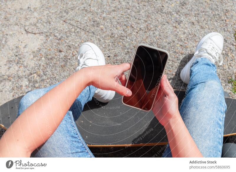 Sitting on skateboard while using smartphone on a sunny day person jeans sneakers outdoor casual technology pavement relaxed sitting hands holding mobile device