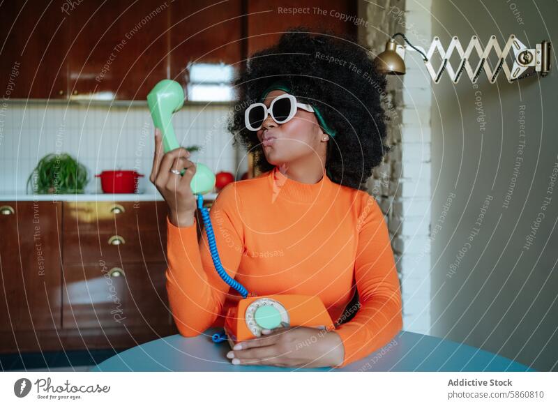 Woman in retro kitchen kissing a vintage telephone woman black 1950s playful afro hairstyle sunglasses styled housewife orange top looking away nostalgic