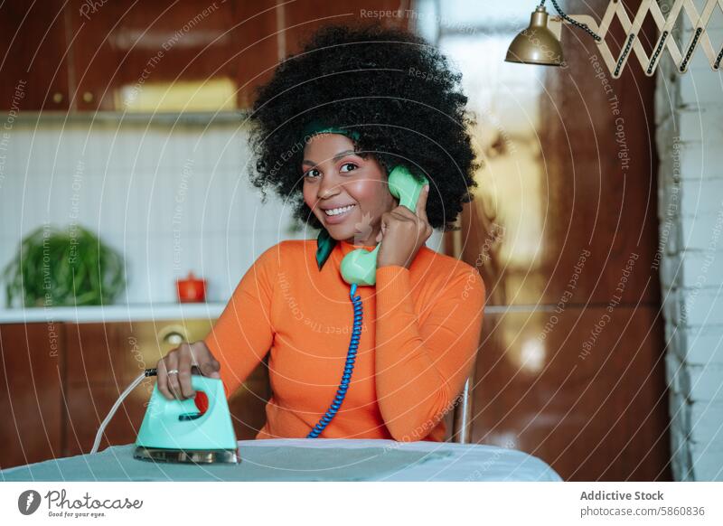 Woman multitasking with vintage phone and iron in kitchen woman black ironing cheerful retro clothes orange shirt telephone hairstyle 50s housewife domestic