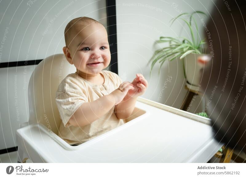 Baby in high chair smiling at father out of frame baby engaging adult playful man warm indoor houseplant cheer clap joy infant white sit interaction happiness