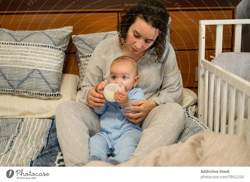 Mother feeding baby in a cozy bedroom setting mother bottle tender care nurturing caucasian pajama comfortable blue pillow decorative crib white warm decoration