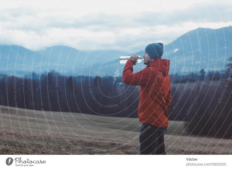 Adventurer on a hike drinks clear water from a glass bottle in a meadow in front of the mountains during a break. Refreshment break male smiling young adult