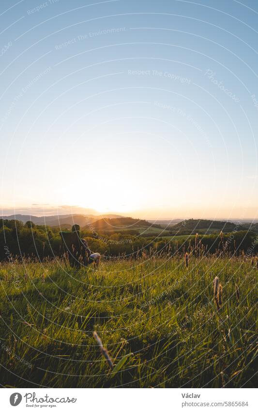 Young man in a checked shirt sits on a light travel chair and enjoys the view of the valley and the setting sun on the horizon. Beskydy Mountains, Czech Republic