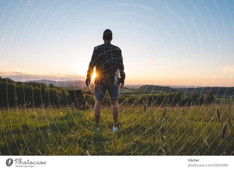 Young man sets up a chair in a meadow at sunset and prepares to relax with a view of the valley and the setting sun on the horizon. Beskydy mountains, Czech Republic