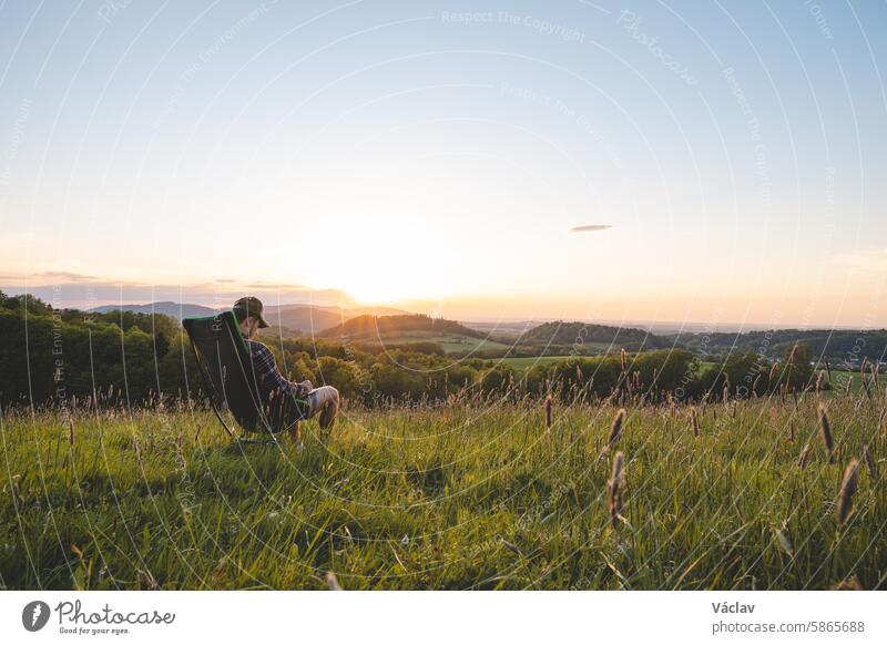 Young man in a plaid shirt sits on a traveling light chair, enjoying the view of the valley and the setting sun on the horizon. Beskydy mountains, Czech Republic