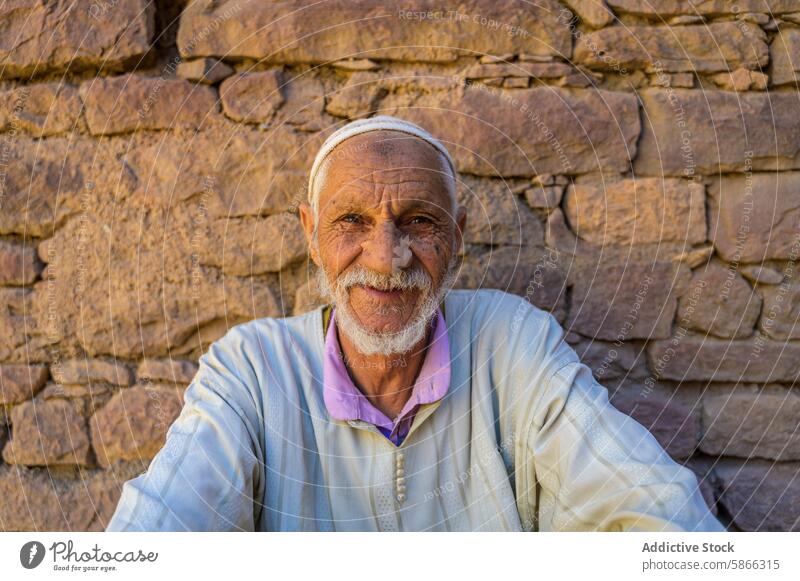 Smiling elderly Moroccan man sitting against stone wall moroccan smile traditional attire hat robe joy senior warm texture north africa happiness portrait