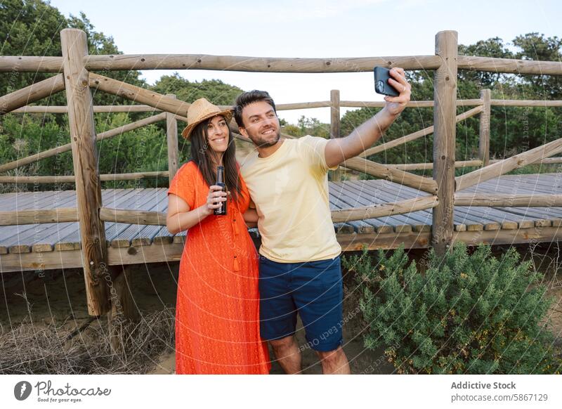 Friends enjoying beers and taking a selfie outdoors friend casual summer woman bridge wooden nature scenery bottle drink mobile phone camera hat smile happiness