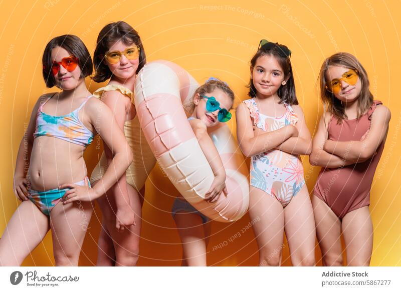 Group of children in swimwear with summer props in studio theme colorful sunglasses pool float joyful vibrant yellow background pose cheerful accessory trendy