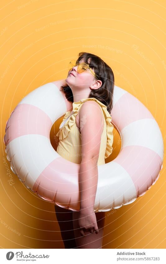 Young girl posing with inflatable swim ring, summer vibe child studio theme yellow backdrop sunglasses pink white stripe outfit vibrant leisure vacation fun