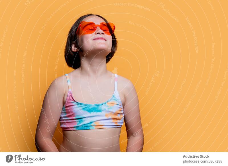 Young girl with heart-shaped sunglasses in summer attire child studio bright orange backdrop sunlight basking tie-dye swimsuit top smile youth cheerful