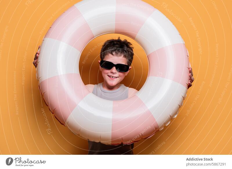 Boy with sunglasses smiling through a swim ring child summer studio theme boy smile orange background happiness joy playful fun leisure vacation youth striped