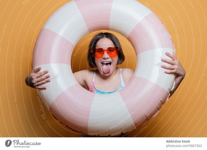 Young girl posing with swim ring in a summer-themed studio child sunglasses tongue playful joy fun vibrant orange background swimwear season happy young kid