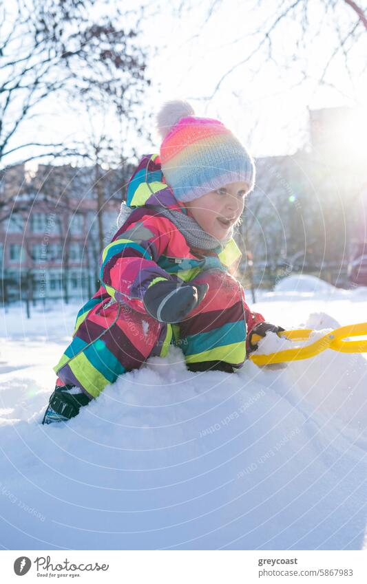 Child in colorful snowsuit playing in snow child park shovel winter outdoor activity trees buildings kid playful young cold environment urban residential city