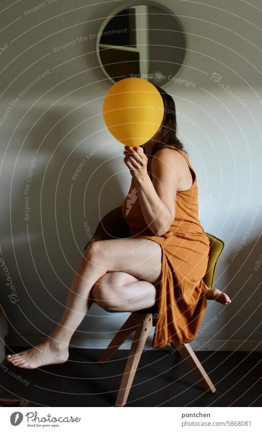 a woman in a dress, sitting on a chair and holding a yellow balloon in front of her face. privacy Anonymous Hide Woman hide sb./sth. secret Safety Face Dress