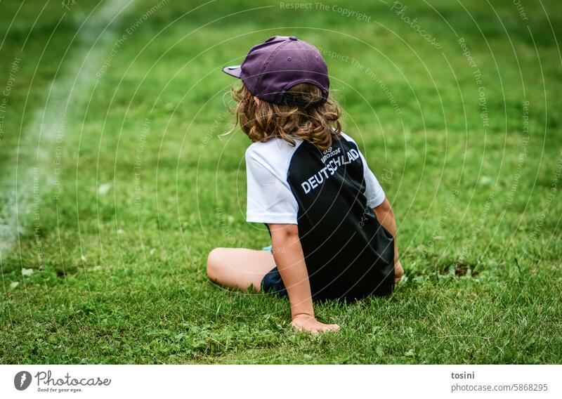 A boy in a soccer jersey takes a break on the pitch Boy (child) Meadow Lawn Child Foot ball Football pitch UEFA European Championship EM2024 Cap Green Sports