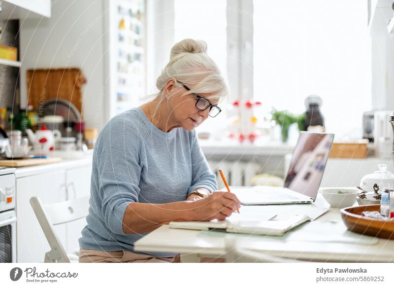 Senior woman with laptop and notebook sitting at the table in the kitchen people senior mature female elderly home house old aging domestic life grandmother