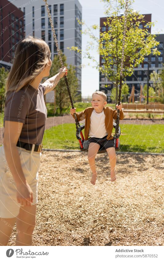 Young boy enjoys swing as mother pushes in park son summer sunny happy family outdoor smiling child woman leisure playtime fun cheerful bonding parent kid