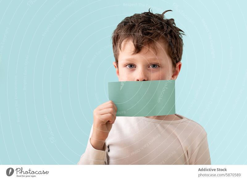 Young boy holding green card over mouth in studio child light beige shirt pale blue background looking at camera covering mouth indoor expression curious