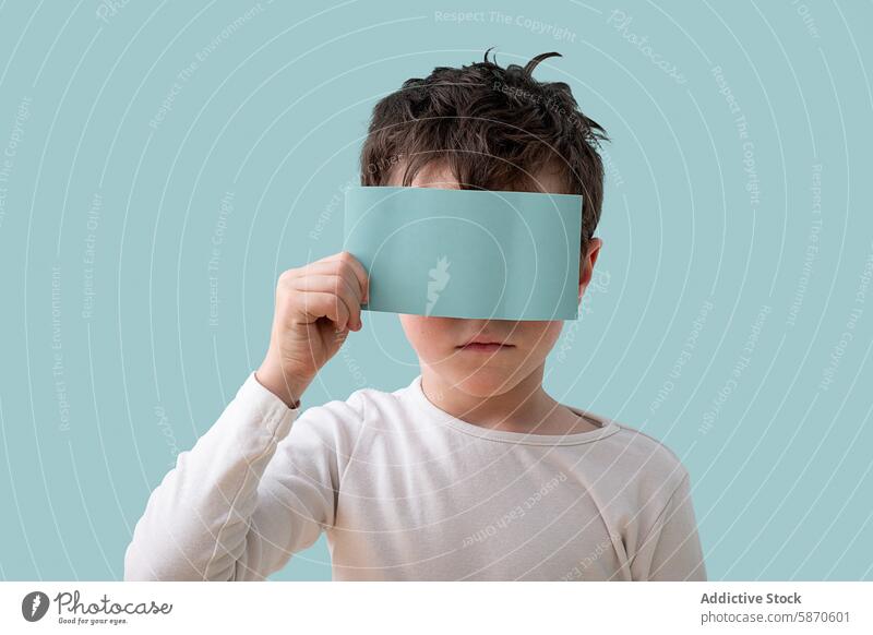 Young boy holding a blue card over his face in studio face covered white shirt caucasian light blue background portrait simplicity expression posture child kid