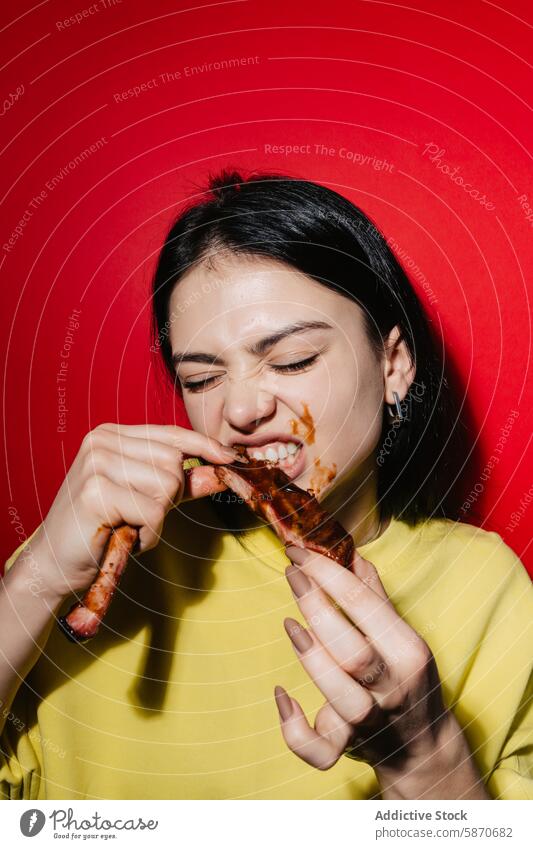 Young woman enjoying messy barbecue ribs against red backdrop eating sauce vibrant yellow shirt food meal enjoyment pleasure youth casual lifestyle dining happy