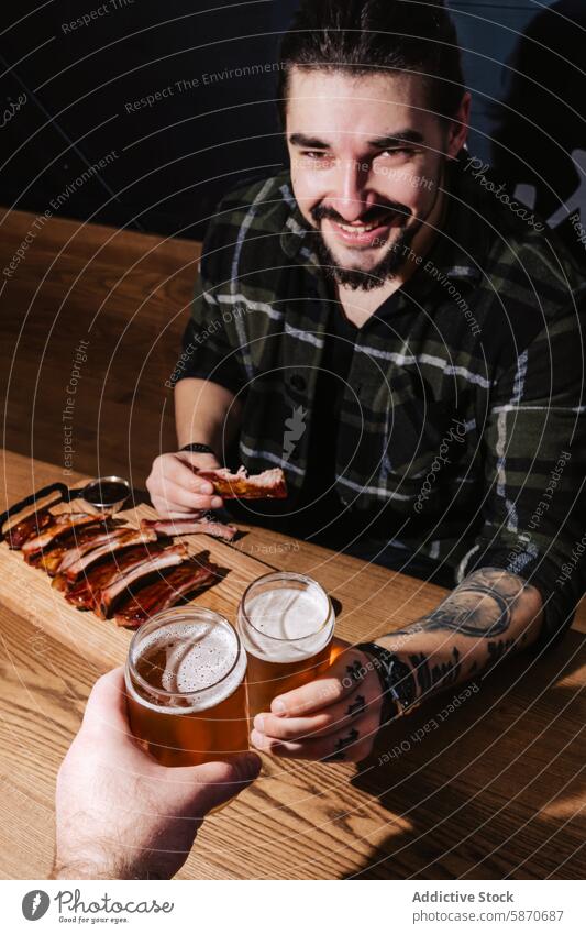 Friendly man enjoys ribs and beer at a barbecue restaurant tattoo beard smile dining friend sharing table wooden glass meal drink laughter pleasure indoor