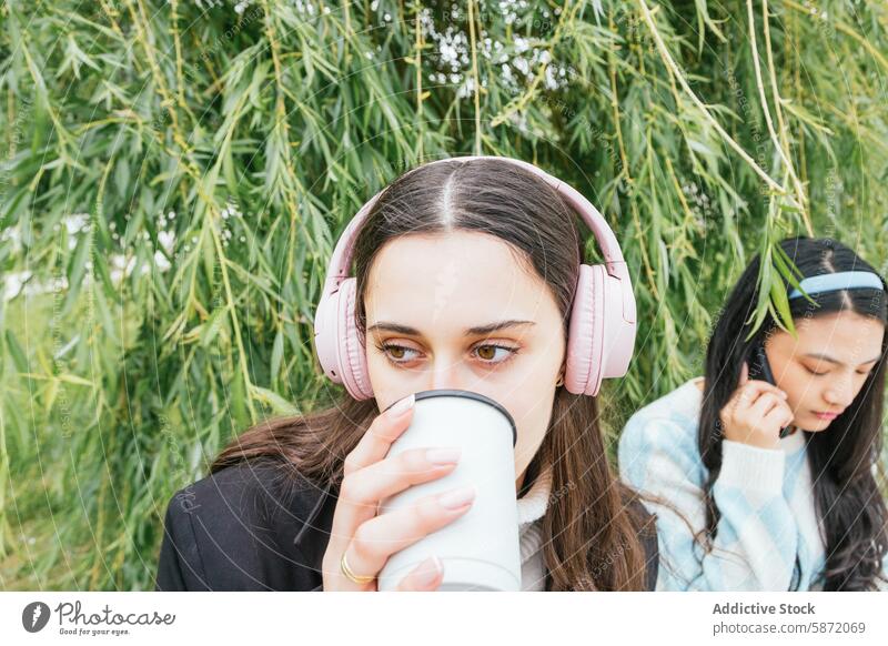 Young students enjoying music and drinks in a nature setting friend headphone outdoor young smartphone woman greenery relax leisure casual youth sip cup bamboo