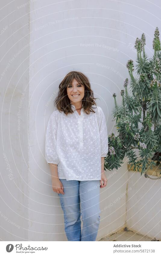 Smiling woman in white blouse stands by a plant smile camera jeans wall green urban tranquil standing cheerful embroidered fashion outdoor daytime casual wear