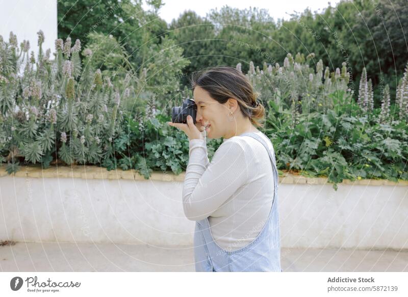 Woman in casual attire photographing garden plants woman camera photography outdoor denim jumpsuit white shirt nature foliage hobby leisure daytime standing