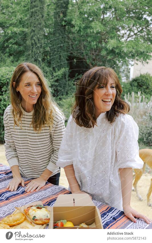 Two women enjoying a picnic outdoors with fresh food adult table conversation joyful lively looking away cloth-covered nature greenery summer casual gathering