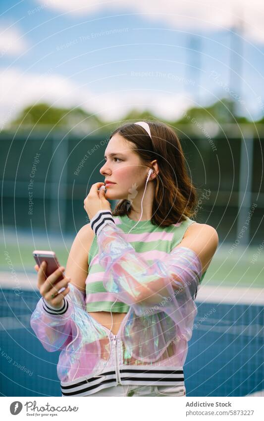 Young woman enjoying music on smartphone at tennis court earphone casual sporty attire young listening holding outdoor leisure activity technology mobile device