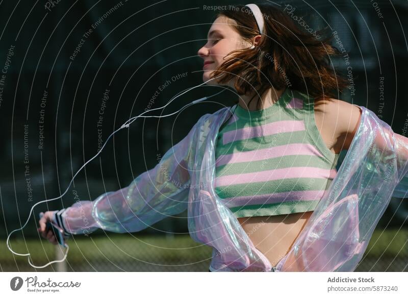 Young woman enjoying music at a sunny tennis court young day listening freedom stylish transparent jacket striped top vibrant bright outdoors leisure relaxation