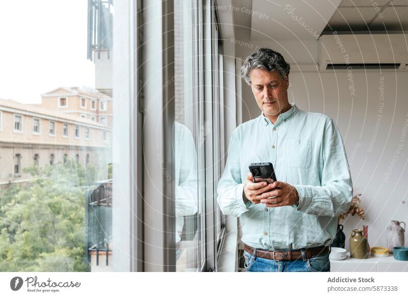 Middle-aged man using smartphone by window in office technology mobile phone gadget device looking down urban architecture modern indoor pastel shirt jeans belt