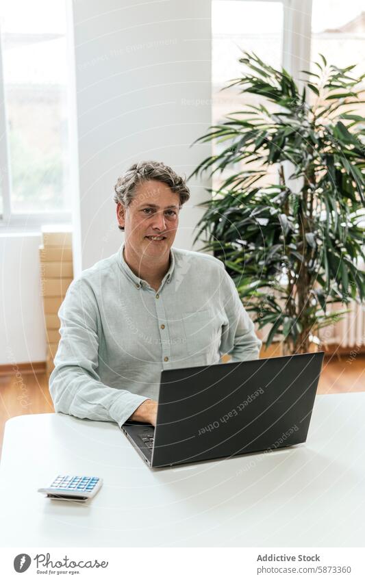 Middle-aged man working on laptop in bright office business technology device gadget indoor desk looking at camera caucasian middle-aged plant calculator mature