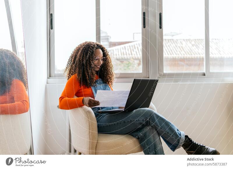 African American woman working on laptop in a bright coworking space african american document armchair natural light window technology device gadget curly hair