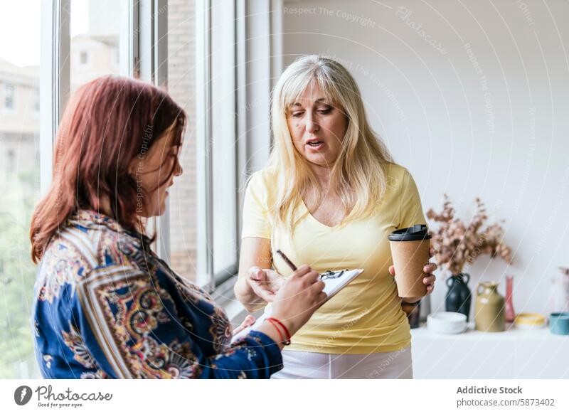 Two women discussing work with coffee and smartphone in office coworking discussion coffee cup clipboard technology business caucasian conversation professional