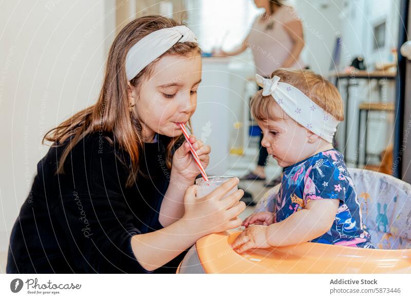 Sisters sharing a drink while mother cooks in the background family home child sister toddler girl cooking kitchen interaction sibling care assistance