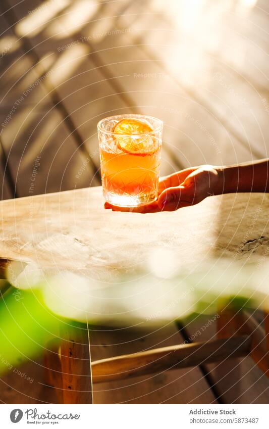 Unrecognizable hand holding a refreshing cocktail at a sunlit outdoor table sunlight summer refreshment orange slice unrecognizable wooden table beverage