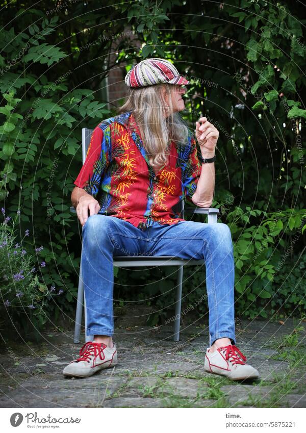 colorful man with cap on a chair in the green Man Long-haired Blonde Chair Sit Cap Hamd jeans variegated sneakers Terrace Nature Profile View to the side