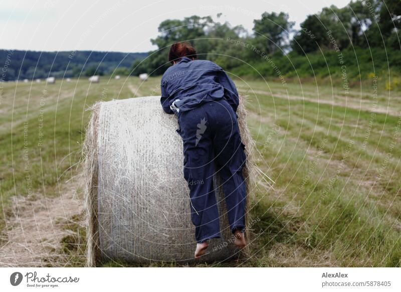 A woman in a kind of boiler suit tries to climb barefoot onto a round bale of straw, you can only see her from behind Landscape Woman Blue overalls afoot