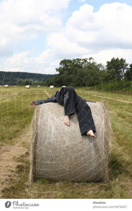 A woman in a kind of boiler suit has climbed barefoot onto a round bale of straw and is now lying on top of it Landscape Woman Blue overalls afoot Bale of straw
