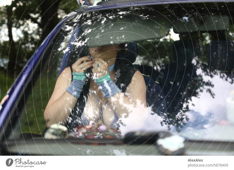 A woman with a large neckline is sitting in the front right-hand side of her car, putting on make-up and jewelry. You can't see her through the reflection in the windshield