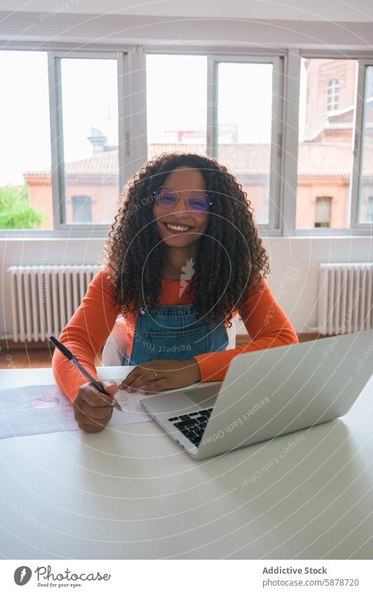 Smiling young woman working in a bright office smile laptop desk business professional glasses curly hair cheerful modern technology workspace entrepreneur