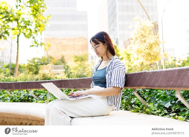 Young woman working outdoors with laptop in urban park seated bench young professional technology greenery building city nature focus casual glasses stylish