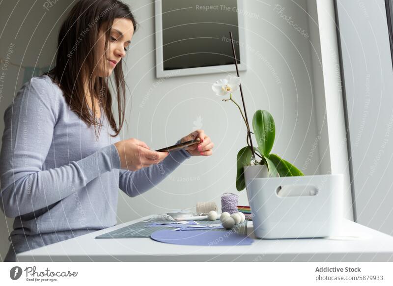 Young woman engaged in paper crafting at home cutting table indoor hobby creative focus concentration scissors supplies smartphone orchid pot simplicity