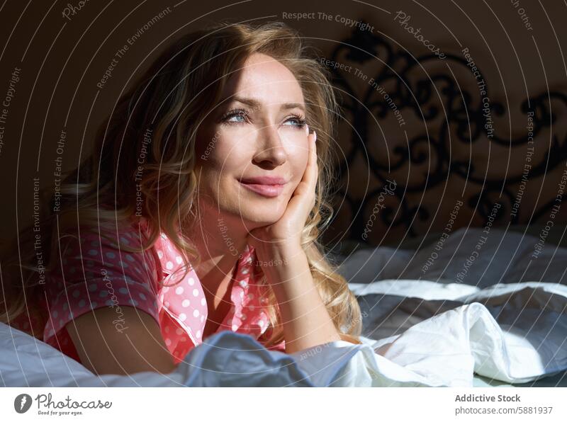 Dreamy blond woman relaxing in bed, illuminated by sunlight morning peaceful serene contemplative gaze expression natural bedroom cozy comfort rest daylight