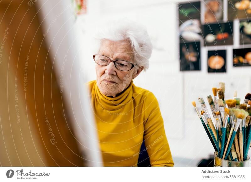 Elderly woman painting in her art studio senior brush creativity elderly mature yellow turtleneck focused hobby glasses workshop canvas experience artistic