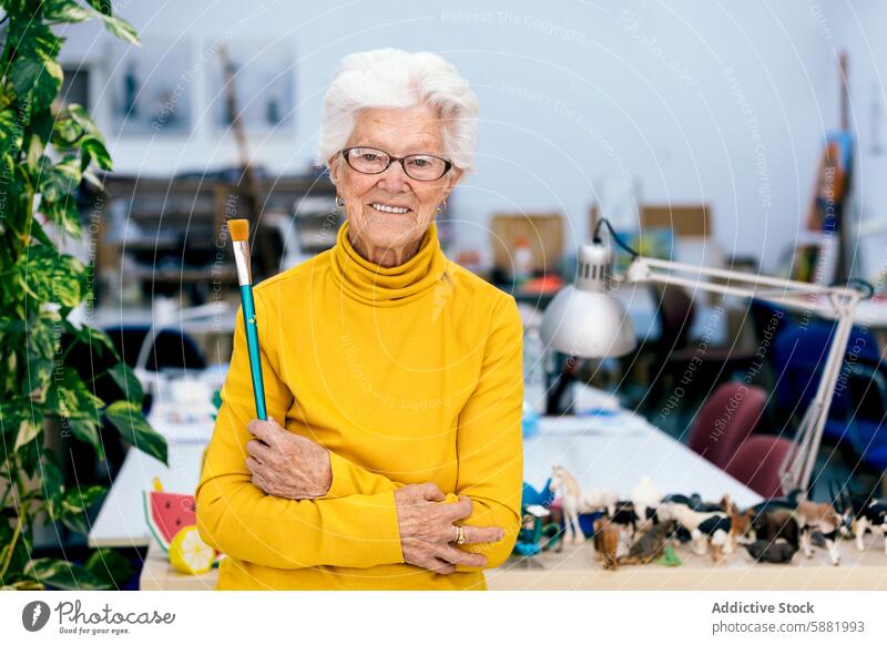 Elderly woman artist holding a paintbrush in studio elderly cheerful senior mature white hair smiling yellow turtleneck arms crossed creativity painter happy