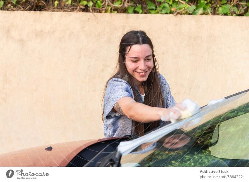 Woman smiling while washing a car outdoors woman car wash windshield cheerful diligent cloth young vehicle car care maintenance manual labor cleaning daytime