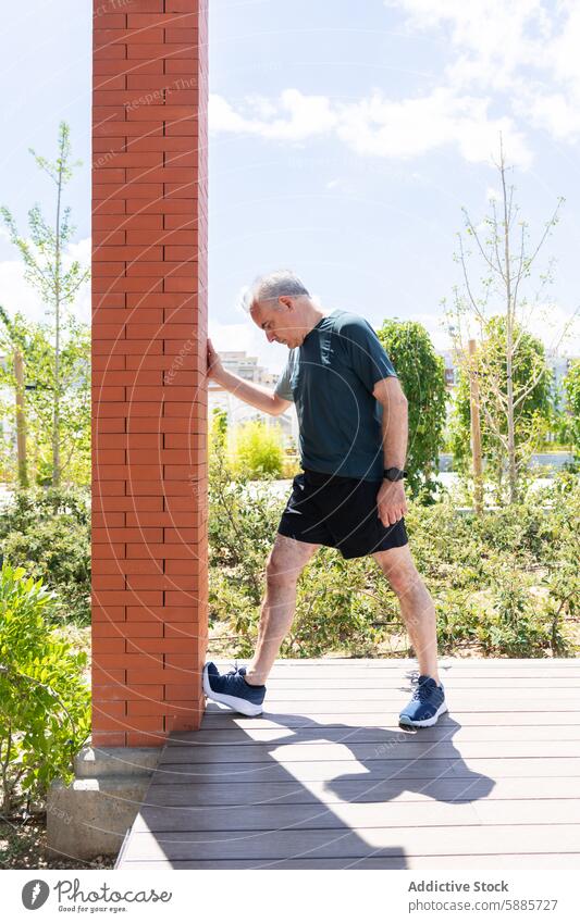 Mature man stretching against a wall in a sunny park senior mature sportswear outdoor leg brick column tree blue sky fitness health wellness exercise activity