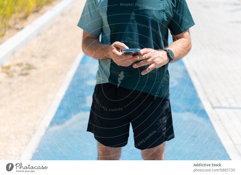 Unrecognizable man checking smartphone while on running track fitness sportswear technology middle-aged exercise outdoor daytime summer athlete health lifestyle